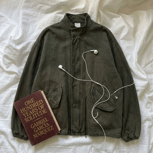 An olive green corduroy jacket is laid flat on white fabric, styled with a book titled 'One Hundred Years of Solitude' by Gabriel García Marquez and white wired earbuds.