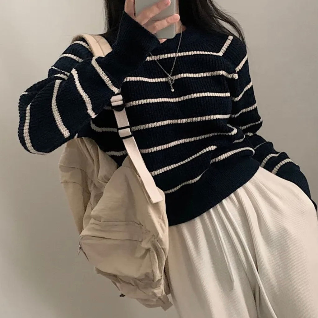 Person wearing a black and white striped sweater with a beige bag and pants against a neutral background